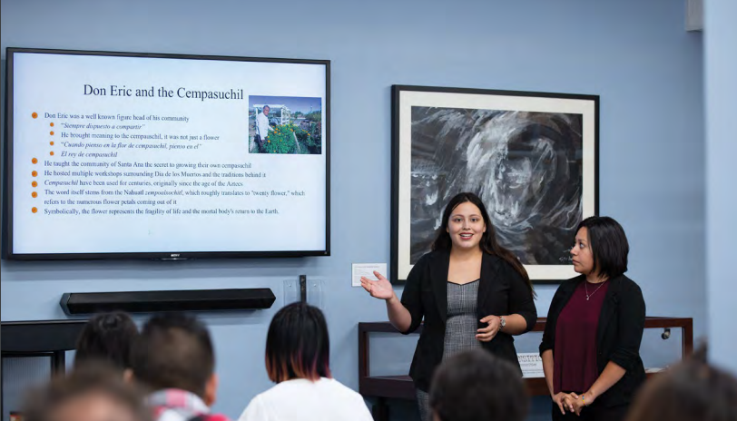 Two women present to an audience about Don Eric and the Cempasuchil using a slideshow on a tv.