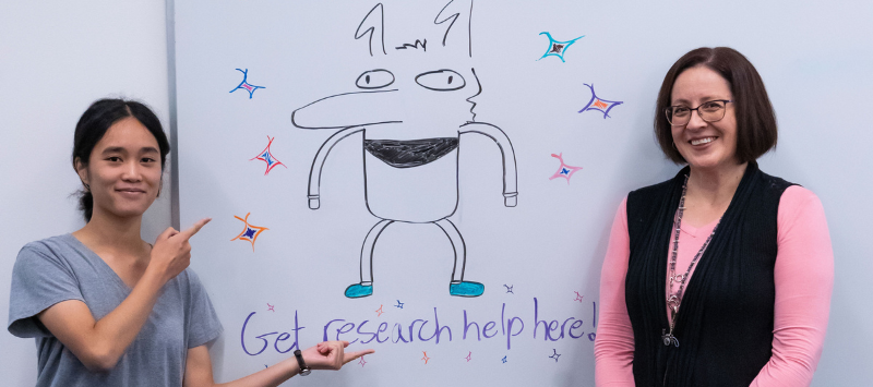 Library Assistant Supervisor Antoinette Avila and a student pose in front of a white board that says "Get research help here!" and features an anteater drawing.
