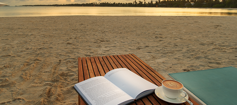 Open book and cup of coffee on a wooden table with sunset beach background.